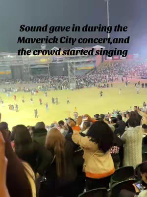 A crowd of people are seen from behind, facing a stage. The stage is lit up with bright lights, and a large crowd of people fill the seats and the open space in front of the stage. Text overlays the image, describing the concert's issue.
