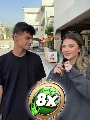 A selfie style shot with a woman holding a microphone, interviewing a man on a street. A coin-like graphic with a green Pepe the Frog stands in the foreground. The man stands on the left, partially in frame, looking at the female interviewer on the right, who is smiling. The background shows a building and a walkway with some open kiosks.