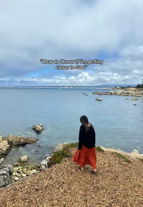 A woman stands on a rocky outcrop near the ocean. She is wearing a black long sleeve shirt and a long orange skirt, viewed from the back. The sky is overcast, and the ocean stretches out in the background. The text overlay is on the sky, and says: “How do I know if I'm getting closer to God”.