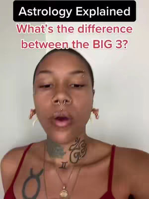 The image focuses on a person, looking directly at the camera with an open mouth, possibly speaking. They have multiple facial piercings, including on the nose and eyebrows. They are wearing a red top, and their neck is covered in tattoos. Overlayed at the top is text in a black box stating "Astrology Explained", followed by another line in pink text saying "What's the difference between the BIG 3?"