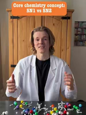 A young man wearing a lab coat stands in front of a wooden cabinet with a table in front of him. On the table are several molecular models. An orange banner at the top says 'Core chemistry concept: SN1 vs SN2.'