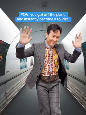 A man in a stylish suit with checkered patterns is depicted in an airport corridor's tunnel, his arms raised with open palms. A light blue text overlay reads "POV: you get off the plane and instantly become a tourist."