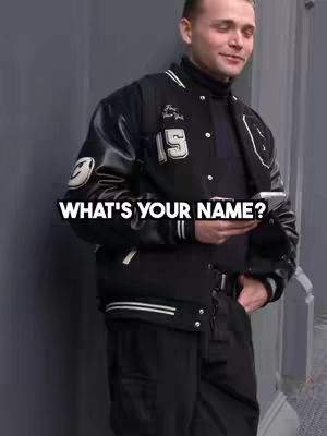 A man in a black and white varsity jacket holds a phone, looking slightly off-camera. Text overlays the image asking 'WHAT'S YOUR NAME?'. The background appears to be a gray wall.