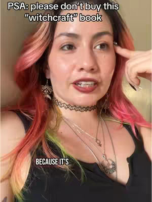 A woman with light skin and pink-orange hair is looking directly at the camera with a slightly concerned expression. She has dark eye makeup, dark lipstick and a few pieces of jewelry. The woman is wearing a black top.