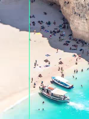 Aerial view of a beach with turquoise water, white sand, and a large cliff on the right side. Boats are docked in the water, and people are scattered on the beach and in the water. A bright turquoise vertical line is on the left side, separating the sand area from the rest.
