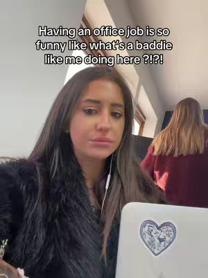 A selfie-style shot featuring a young woman in a fur-like coat, seated in an office setting. She's the primary focus, looking directly at the camera with a slightly quizzical expression. Behind her, blurred out, is another person, adding depth to the scene. The upper portion of an Apple laptop with a heart-shaped sticker is visible in the foreground. Text overlay dominates the top of the frame with white text set against the visible setting.