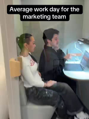 A scene inside what appears to be an office or meeting space, with two people seated. A woman with green-tinged hair, wearing a white top and dark pants, is seen in the side profile, sitting on the toilet and looking towards the left. Next to her sits a man, seen pointing towards a laptop screen to the right. The setting is enclosed by transparent glass, suggesting a small, private office arrangement. The lighting is bright, and the overall composition is a documentary-style.