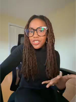 A woman with long, brown dreadlocks wearing a black turtleneck and large-framed glasses with tortoise shell and white accents is looking directly at the camera while holding out her right hand. She is seated in a chair in what appears to be a home office, with a window and door in the background.