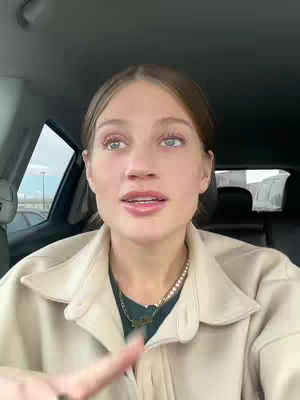 A woman with light skin is inside a car, looking at the camera and talking. She has light-brown hair and is wearing a light-colored jacket and a necklace. The background shows a blurred view of the outside, suggestive of a parking lot. The camera is positioned in front of the woman, capturing a close-up shot.