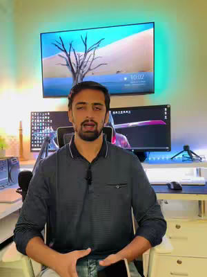 A man with a beard is seated in front of a desk setup, speaking to the camera. Behind him are two computer screens, and a large TV screen displays an image of a tree. The room is lit with a combination of natural and artificial light.