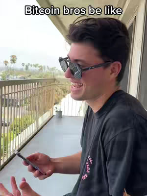 A man wearing sunglasses and a black t-shirt holds a phone on a balcony. Text overlay says 'Bitcoin bros be like'. The scene shows an outdoor setting with a railing, trees, and a building in the background.