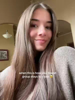 A selfie-style shot of a young woman smiling, framed from the chest up. She has long, wavy brown hair and wear a light-colored sweater. The background is slightly blurred, showing a warm-lit interior setting. Text overlayed on the image reads 'when this is how your friend group stays so close'. The composition is intimate and friendly, with a focus on the woman's expression.