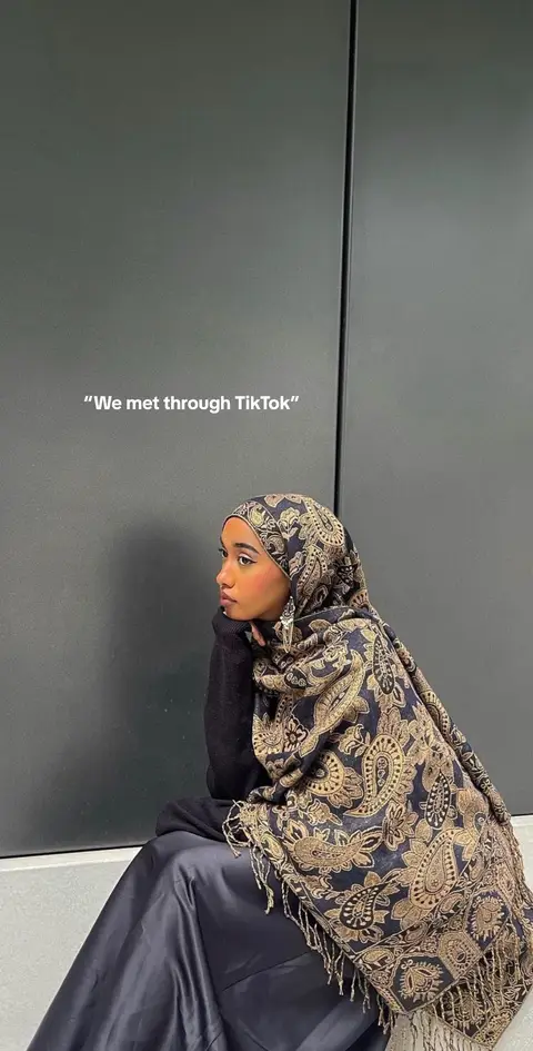 A woman wearing a patterned headscarf and dark clothing leans against a dark gray wall, resting her chin on her hand and looking to the side. Text overlayed on the image reads, "We met through TikTok".