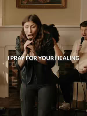 The image shows a woman speaking into a microphone, hands clasped near her chest. She is surrounded by others, and overlayed text reads 'I pray for your healing.' The setting appears to be a music performance venue.