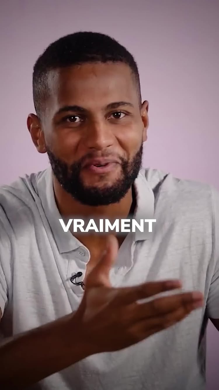 A medium shot showcasing a man with short, dark hair and a trimmed beard. He wears a light gray polo shirt and is positioned against a soft, pink background. The word 'VRAIMENT' is superimposed in white, bold text across his chest.