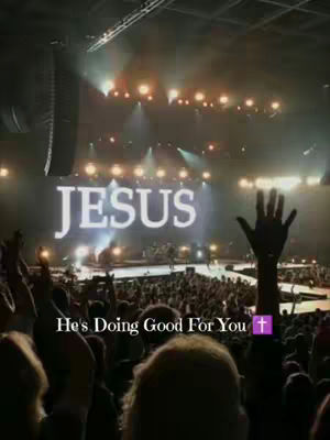 A large crowd of people with their hands raised, facing a stage with a screen that says "JESUS." The stage is lit with bright lights, and there is a text overlay at the bottom that says, "He's Doing Good For You," with a purple cross icon.