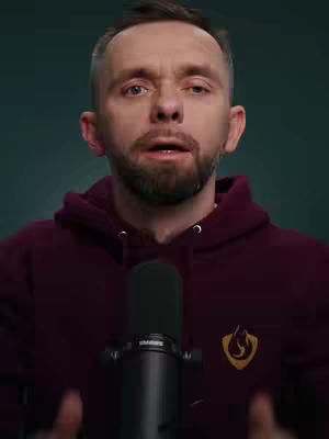 A medium shot shows a man with short, dark hair and a beard, wearing a burgundy hoodie. He is positioned in front of a dark background and behind a black microphone. The man's face is expressive, and the overall composition suggests a focus on communication or commentary.