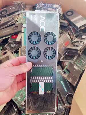 The image shows a close-up of a hand holding a large black and silver computer component, possibly a graphics card. The component has multiple cooling fans and various electronic components visible. It is surrounded by a pile of other similar discarded computer parts, suggesting a recycling or repair context.