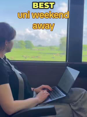 A woman is sitting on a bus, looking out a window at a green landscape. She is using a laptop. Text overlays say "BEST uni weekend away".
