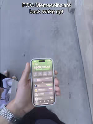 A first-person perspective showcasing an open phone with an app interface displaying financial data. The phone, held against a gray background, likely a sidewalk, shows a balance of $308,591.47. Text overlayed at the top reads, 'POV: Memecoins are back wake up!' setting the context for the crypto-related content.