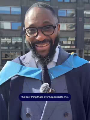 A smiling man in graduation attire, wearing glasses. He is outdoors with a building visible in the background. An overlay text bubble at the bottom says “the best thing that’s ever happened to me.”