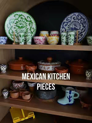 A well-lit shot showcases a variety of Mexican ceramic pieces artfully arranged on wooden shelves. The shelves hold a range of colorful plates, bowls, cups, and serving dishes, creating a vibrant display of traditional craftsmanship. The composition is a still life with a focus on showcasing the collection within a cozy, indoor setting.