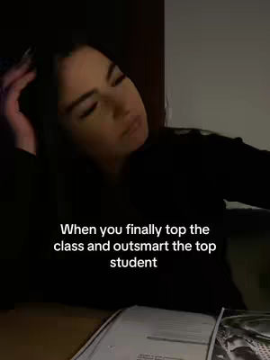 A woman in a dark-colored top is resting her head on her left hand, with eyes closed. In the foreground, there is a desk with papers. The background is dimly lit.