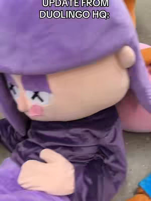 The image shows the Duolingo mascot, an owl dressed in a purple outfit with cross marks on its eyes, suggesting it's deceased, under a blurred background.