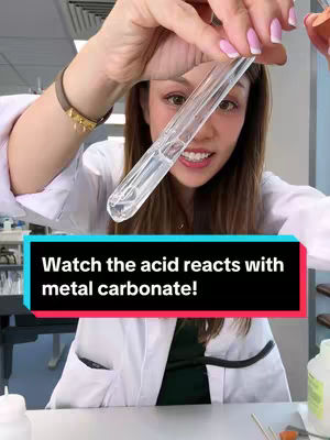A woman in a lab coat holds a test tube with liquid, smiling at the camera. The background suggests a science lab setting.