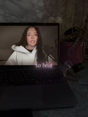 A laptop is open, displaying a woman on the screen who is speaking. The text overlay "to heal" is present on the screen image. The laptop is on a desk next to items that are not clearly visible.