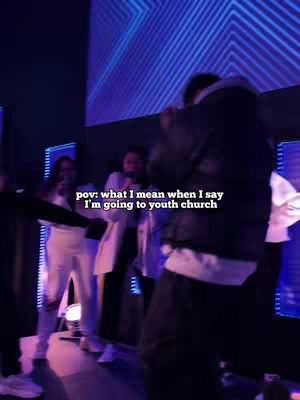 The image is a low-lit, slightly blurred video still taken from a youth church service. The primary focus is on a person holding a microphone and singing or speaking passionately, their back mostly to the camera. They are wearing a dark jacket and dark pants with a white shirt. Behind them, several other young people are visible, some holding microphones and appearing to sing as well. The backdrop is a large screen showing blue flowing lights or patterns. The overall composition suggests a live music performance or a worship service, with a focus on energetic participation.