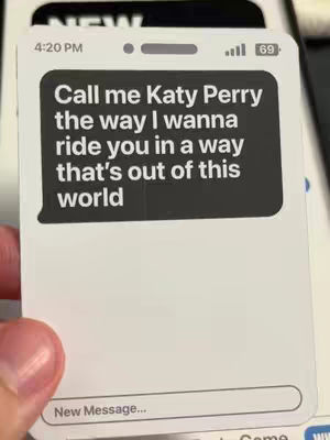 A close-up shot of a smartphone screen displaying a fake text message. The message bubble is a dark gray, containing the words: 'Call me Katy Perry the way I wanna ride you in a way that's out of this world.' The background is blurry, suggesting the phone rests on a surface with a hand holding the phone. The overall composition is intimate and suggestive.