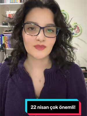 A woman with dark wavy hair and glasses is centered in the frame. She's wearing a purple sweater. Behind her are shelves with books and decorative items. A text overlay at the bottom says, "22 nisan çok önemli!".