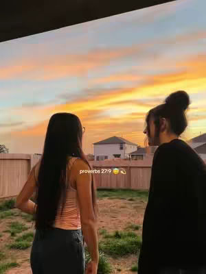 Two women stand side-by-side, seen from behind, framed against a vibrant sunset. The sky is a gradient of oranges, yellows, and pinks. They are in a backyard setting, with a wooden fence and a house visible in the background. The text overlay says 'proverbs 27:9'.