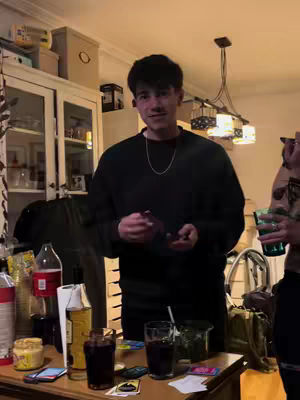A man is standing in a room, smiling while holding an object in his hands. Behind him, a table is set with various items, including alcohol bottles and glasses, suggesting a social gathering. The background shows a cabinet, adding a domestic touch to the scene.