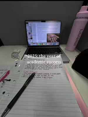 A laptop displaying a document with a hand image is in the center towards the back, with a notebook and pen in the foreground. A pink water bottle is on the right. Text overlay announces the year of academic success.