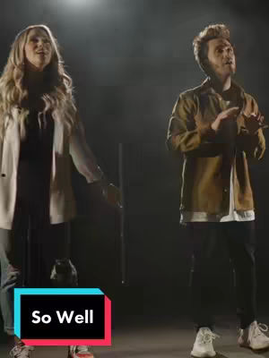 A cover image featuring a man and a woman in a performance setting. They're both looking upwards. The woman is on the left, dressed in black, under a cream open jacket, and holding her arms out. The man on the right wears a brown jacket and black pants, holding his hands up as well. Text in a colored box at the bottom reads 'So Well'. The background is dark, with lighting or spotlight effects. The color palette is muted and clean.
