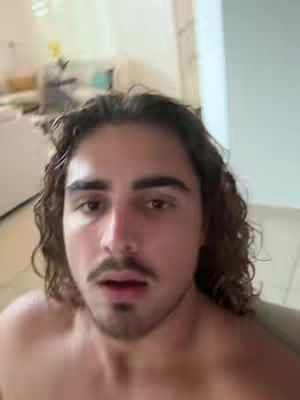 A man with long, wavy brown hair and a mustache looks directly at the camera, his expression open. The background is a blurred domestic interior, suggesting closeness.