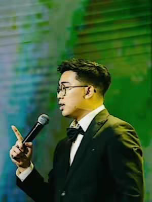 A man in a suit and bow tie speaks into a microphone in front of a blurred green and blue background.