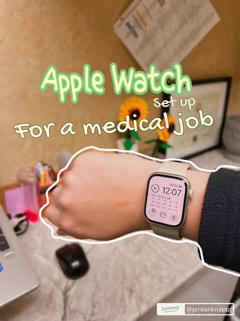 A close-up shot of a person's wrist wearing an Apple Watch, with the watch face prominently displaying the time (12:07) and the date (Mon 30), a few icons, and pink accent colors. The person's arm is positioned over a light-colored surface, and the background includes a blurry desk setup.