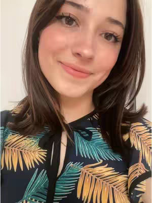 A close-up selfie shows a young woman with brown hair and light skin, smiling gently at the camera. Her cheeks are subtly flushed, her lips are colored a soft pink, and her eyes have a light makeup. Her hair is parted on the side and frames her face. She wears a dark shirt with a colorful tropical leaf design. The background is plain and out of focus.