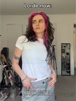 A person with pink-streaked hair is standing in a room, hands in pockets, looking away from the camera. Text overlayed on top of the image indicates who is the topic of the video. The background shows a sparsely furnished room with a large mirror, tripod, and other objects.