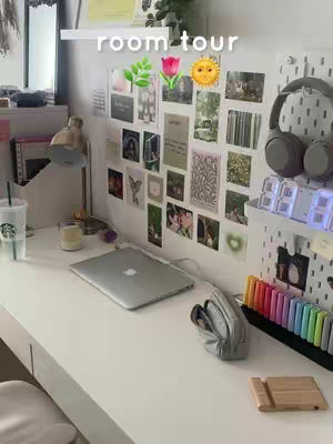 A well-lit, aesthetically pleasing room scene featuring a neatly organized desk. The desk is white and cluttered with various items, including a laptop, a Starbucks cup, a desk lamp, a candle, colored markers, and other decorative elements. The background is a white wall adorned with photos, prints, and decorative items. The overall composition suggests a study space.