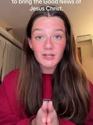 A young woman with long brown hair and fair skin is looking directly at the camera with a slight smile. She is wearing a red top, and her hands are clasped together in front of her chest. The background is slightly blurred, suggesting an indoor setting.