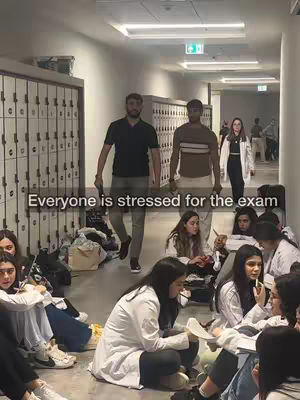A photo taken in a university hallway shows students sitting on the floor and a few people walking. The hallway is lit by fluorescent lights, with lockers on one side. The students on the floor appear to be studying. The lighting suggests an indoor environment, and the overall mood seems to be related to studying or exams.