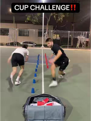 Two men are shown in a competition setting, on a court with lines and a bag in the foreground. Blue and red cups are used in the challenge.