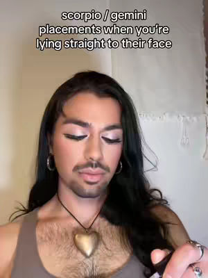 A close-up shot features a person with long black hair, wearing makeup, and a beard. The image prominently displays text at the top, with a background that looks like a neutral-colored wall or curtain. The person is accessorized with hoop earrings and a heart pendant necklace.