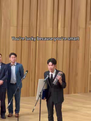 The image features a man in a suit standing on a stage holding an award and a certificate. He is speaking into a microphone, and there are other people in suits standing behind him. The background is a plain wooden wall. Text overlay reads: You're lucky because you're smart