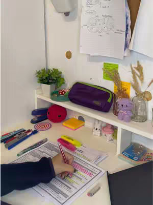 The image is of a desk setup for studying, with a hand holding a pen over a paper. Various stationery items and decorations are on the desk. The color scheme is bright with a mix of colors.