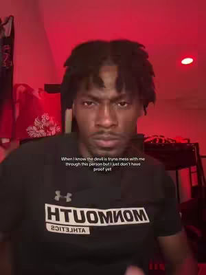A selfie-style video cover image features a young Black man with short dreadlocks, looking towards the camera with a serious expression. He's wearing a black t-shirt with white text that is partially obscured, displaying upside-down letters. The background is dimly lit, with red hues and shadows suggesting an indoor setting. Text overlayed on the image displays the phrase "When I know the devil is tryna mess with me through this person but I just don't have proof yet".